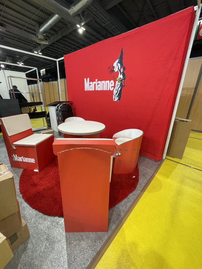 Aménagement du stand du Journal Marianne avec des tables et des chaises en carton