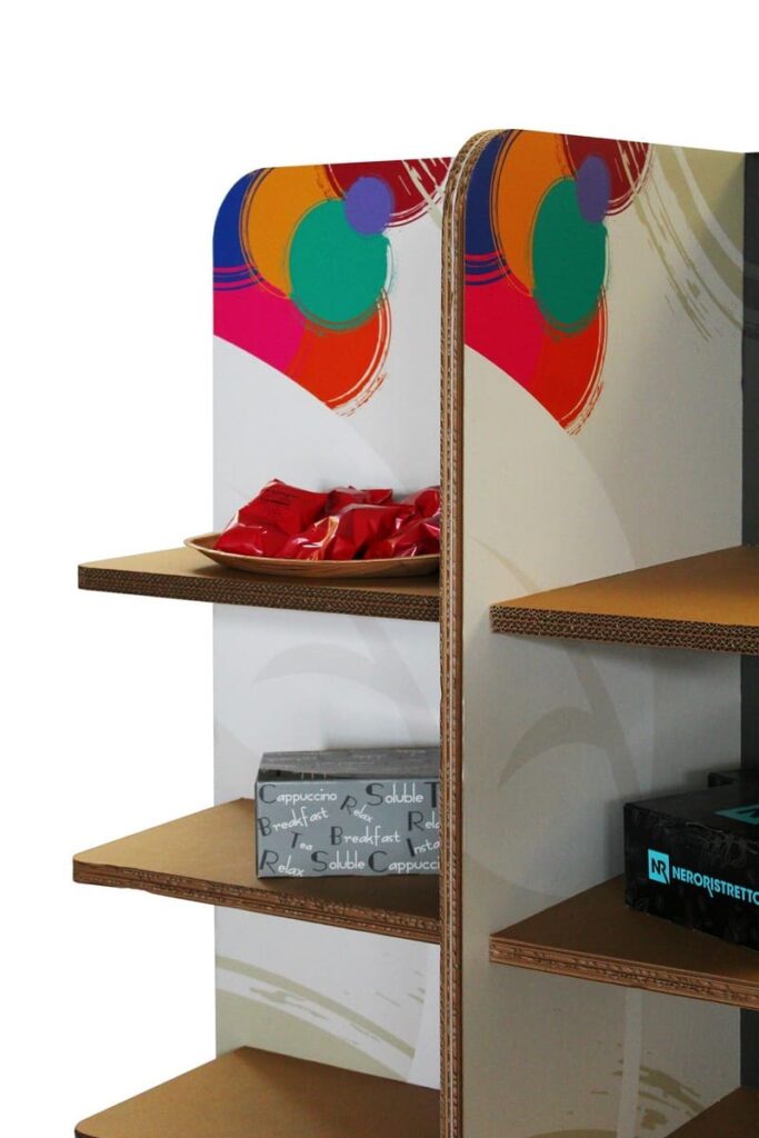 Présentoir en carton coloré et design pour l'aménagement de magasins, avec plateau supérieur décoré d'éléments graphiques vibrants et étagères inférieures organisant des produits café et boissons (café soluble, cappuccino, expresso) dans un espace commercial esthétique et fonctionnel - Le Meuble en Carton une marque du groupe Natur Design