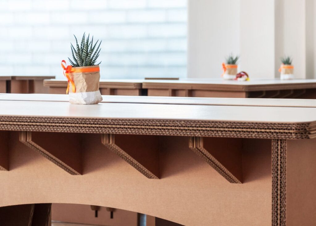 Bureau en carton écologique avec plateau robuste et structure en panneaux cartonnés, intégré dans un espace d'aménagement professionnel pour entreprises ou salles de pause. Décoration végétale discrète avec pot en tissu réutilisable et plante verte, reflétant une approche durable et moderne du mobilier d'office - Le Meuble en Carton une marque du groupe Natur Design