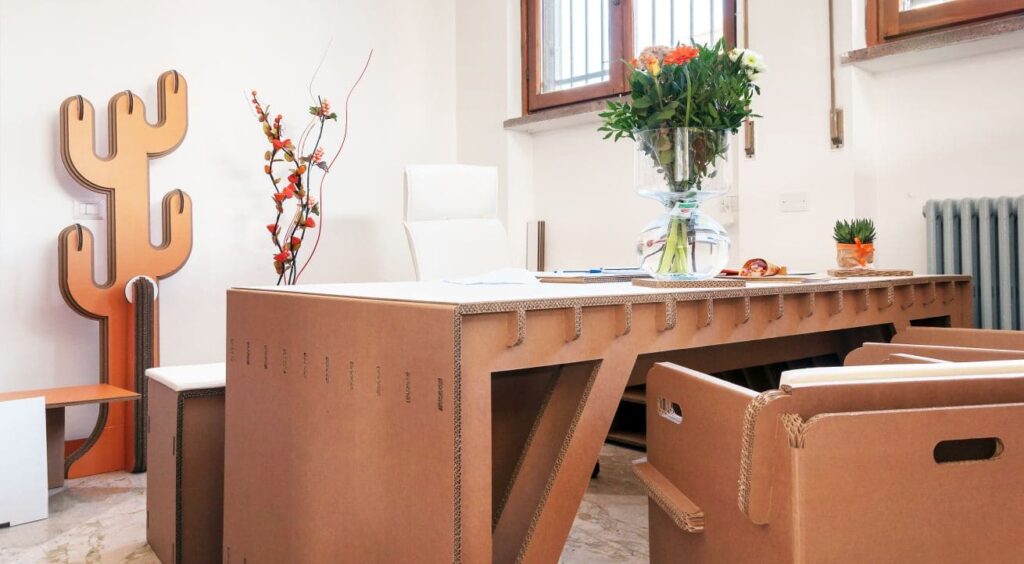 Bureau en carton KARISIMBI avec plateau visible, intégré dans l'aménagement d'entreprise ou salle de pause moderne. Structure cartonnée renforcée pour un mobilier écologique et durable, idéalement positionné pour les espaces collaboratifs et créatifs. Décoration naturelle avec vase floral et éléments organiques (poignées métalliques visibles) qui valorisent le cadre professionnel - Le Meuble en Carton une marque du groupe Natur Design
