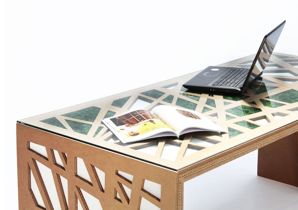 Bureau en carton écologique avec plateau transparent décoré d'un motif géométrique floral, conçu pour les espaces professionnels comme les bureaux et salles de pause. Affichage d'un ordinateur portable ouvert et d'un magazine ouvert sur la surface, illustrant son usage polyvalent dans un environnement moderne et durable - Le Meuble en Carton une marque du groupe Natur Design