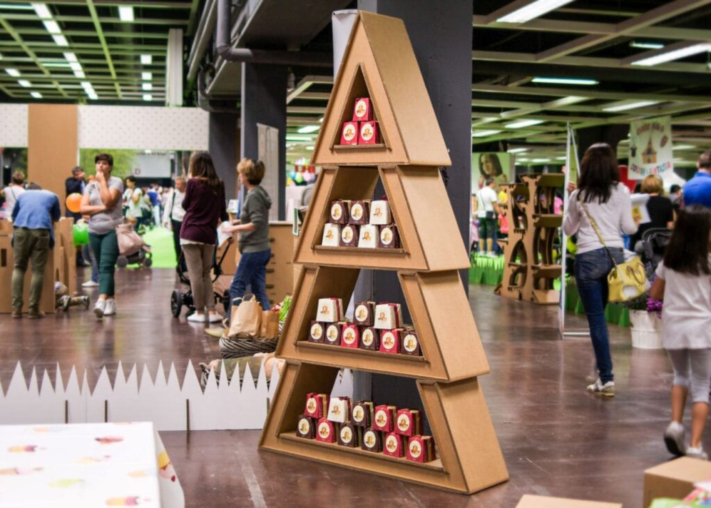Présentoir en carton triangulaire NANDA pour stand événementiel ou décor scénographique, rempli de produits personnalisés et exposés en hauteur dans un espace dédié aux événements professionnels comme les salons ou stands en carton. Structure modulable idéale pour l'aménagement de magasins ou la scénographie d'expositions - Le Meuble en Carton une marque du groupe Natur Design