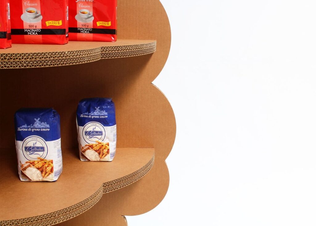 Présentoir en carton KAILA pour l'aménagement de magasins, support structuré en panneaux cartonnés avec étagères perforées et plateau arrondi. Disposé pour exposer des produits alimentaires comme des sachets de café Magnitino et de farine Bolbake dans un environnement professionnel - Le Meuble en Carton une marque du groupe Natur Design