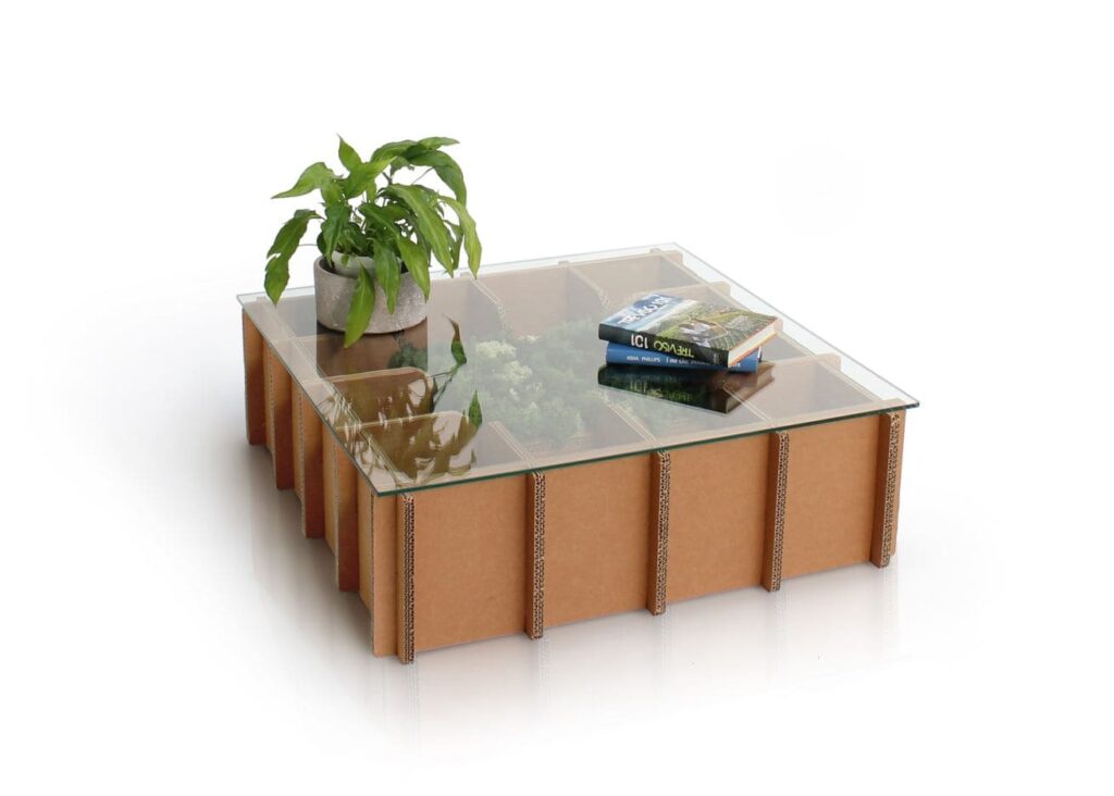 Table basse en carton écologique avec plateau transparent et compartiments intégrés, idéale pour les espaces professionnels ou salles de pause. Structure cartonnée renforcée aux angles, décorée d'un vase contenant une plante verte et d'une pile de livres sur le plateau central - Le Meuble en Carton une marque du groupe Natur Design