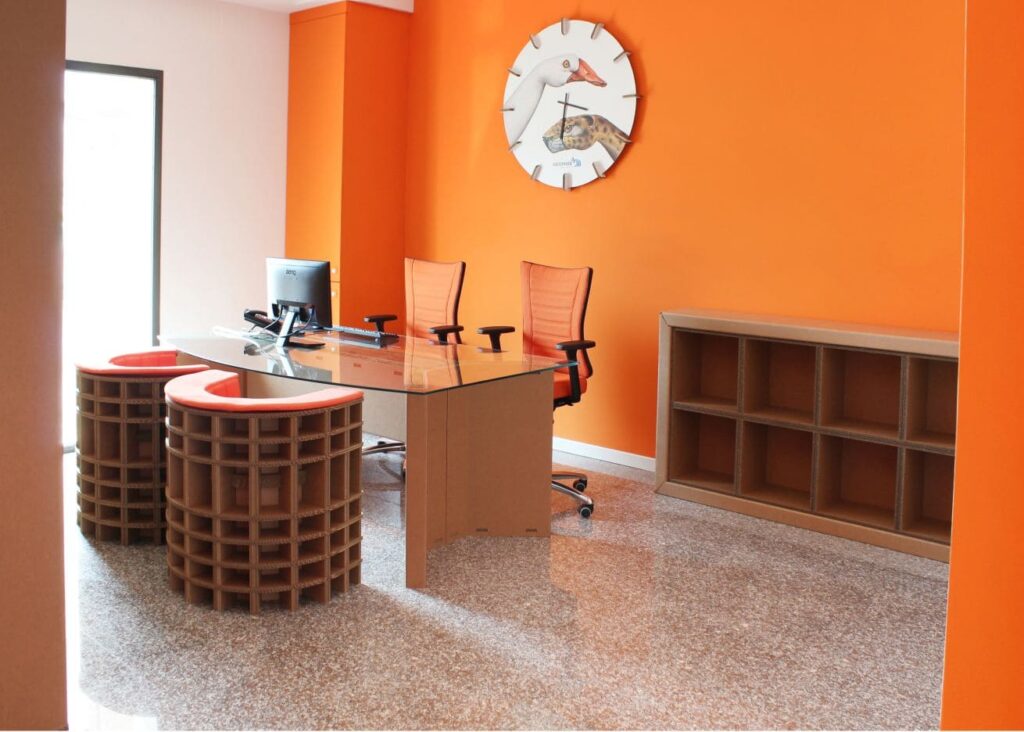 Table de réunion cartonnée moderne pour 6 ou 8 personnes, plateau transparent intégré avec accoudoirs ergonomiques et pieds stabilisés, combinée à des chaises haut de gamme dans une salle de pause professionnelle ou bureau d'entreprise, avec rangements muraux en carton et accessoires design comme coussins colorés et support mural créatif - Le Meuble en Carton une marque du groupe Natur Design