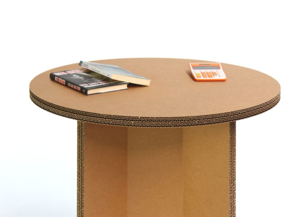 Table en carton rond avec plateau structuré en couches de papier recyclé, idéale pour les espaces professionnels comme les bureaux et salles de pause ou les stands événementiels. Visible sur la surface plate une pile d'ouvrages et un appareil électronique, illustrant son usage polyvalent dans des environnements écologiques et durables - Le Meuble en Carton une marque du groupe Natur Design