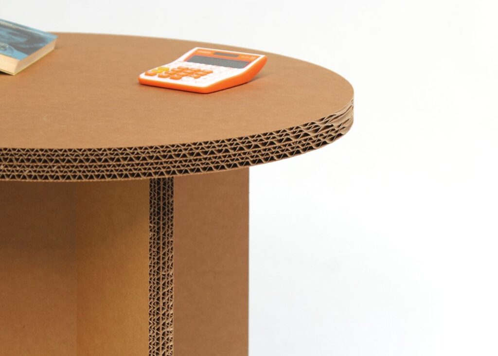 Table en carton design pour espaces professionnels et événements : plateau arrondi et structure robuste en carton recyclé, idéale pour les bureaux, salles de pause ou stands événementiels. Structure stable avec finition ondulée visible sur les côtés - Le Meuble en Carton une marque du groupe Natur Design