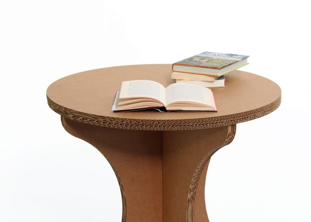 Guéridon cartonné TEBOULO avec plateau texturé en carton ondulé, idéal pour les espaces professionnels comme les salles de pause ou événements scéniques. Montage visible sur une surface plate avec des livres ouverts en guise d'exemple d'utilisation dans un cadre écologique et durable - Le Meuble en Carton une marque du groupe Natur Design