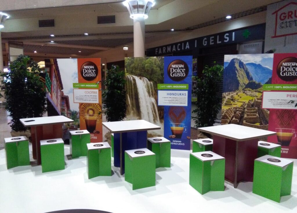 Tabourets en carton écologiques pour stands événementiels professionnels avec plateaux intégrés, positionnés autour de tables et affiches marketing Nescafé Dolce Gusto sur des décors thématiques café, dans une scénographie salonnée pour salons et espaces commerciaux - Le Meuble en Carton une marque du groupe Natur Design